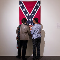 Two people standing in front of a confederate flag that is unraveling