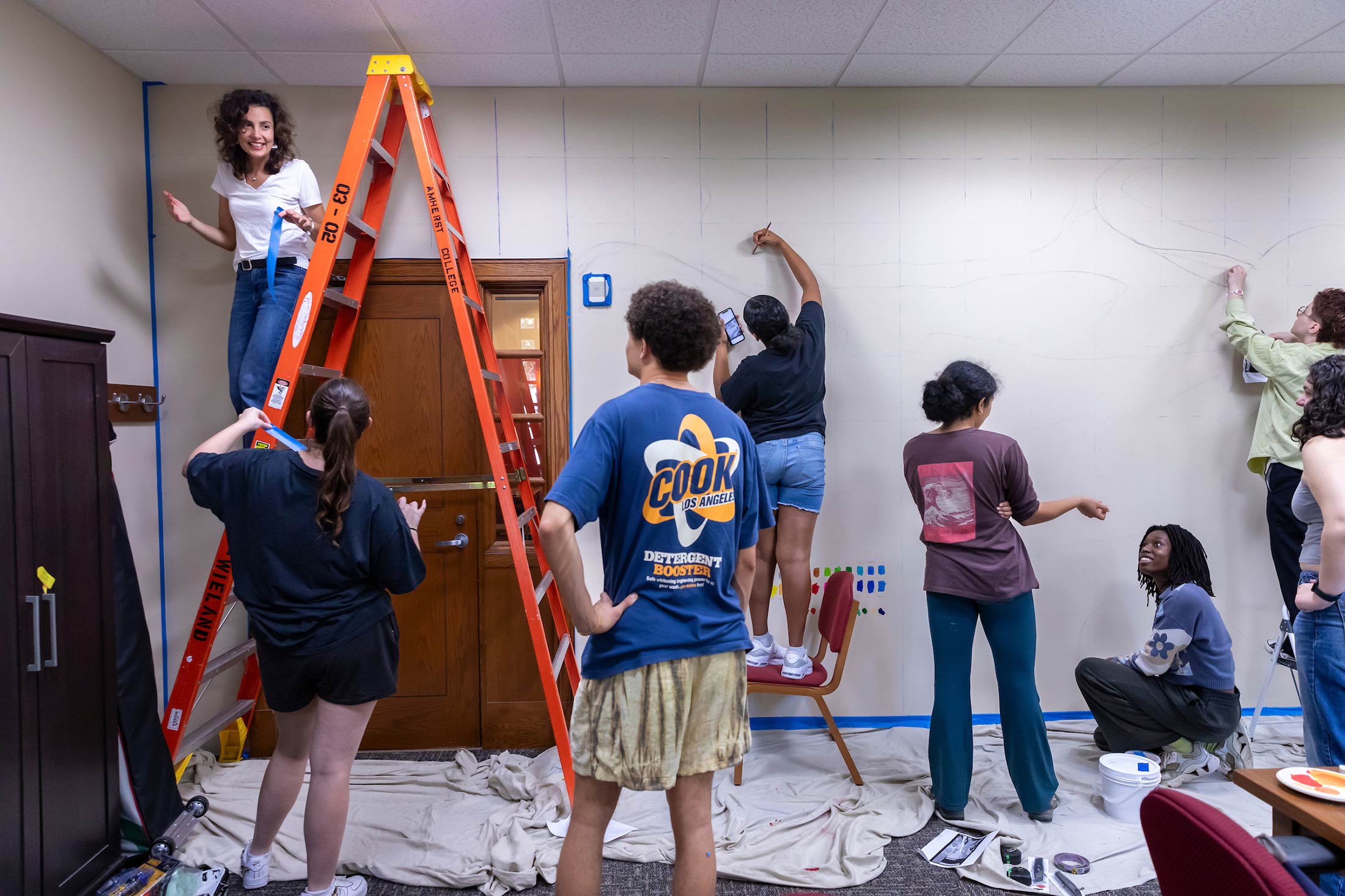 Art students, one on a ladder, begin painting a mural on a wall.