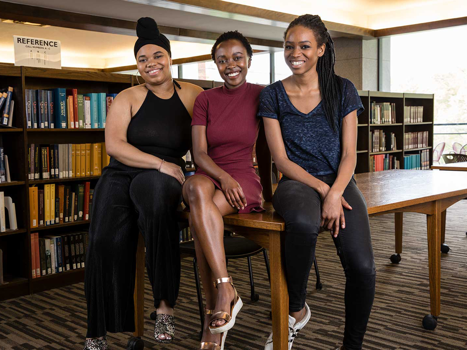 Three Black woman sitting on a table in a library smiling
