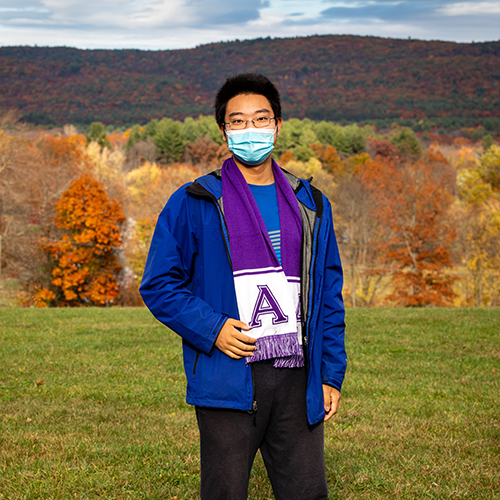 A young student in a mask standing in front of fall foliage