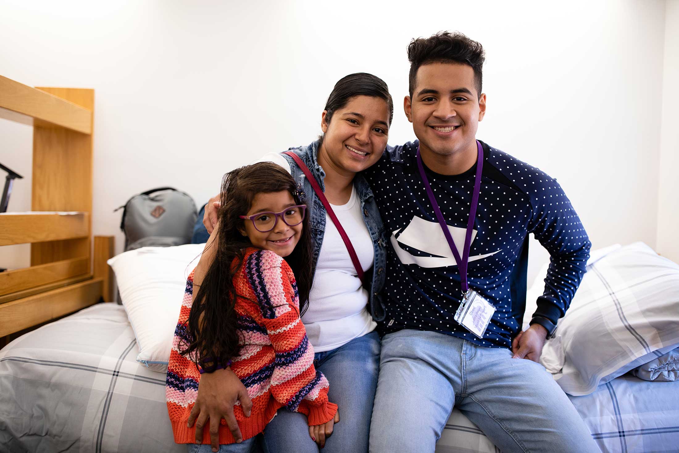 A first year student sitting on a bed in his new residence hall, with his arms around his mother and little sister