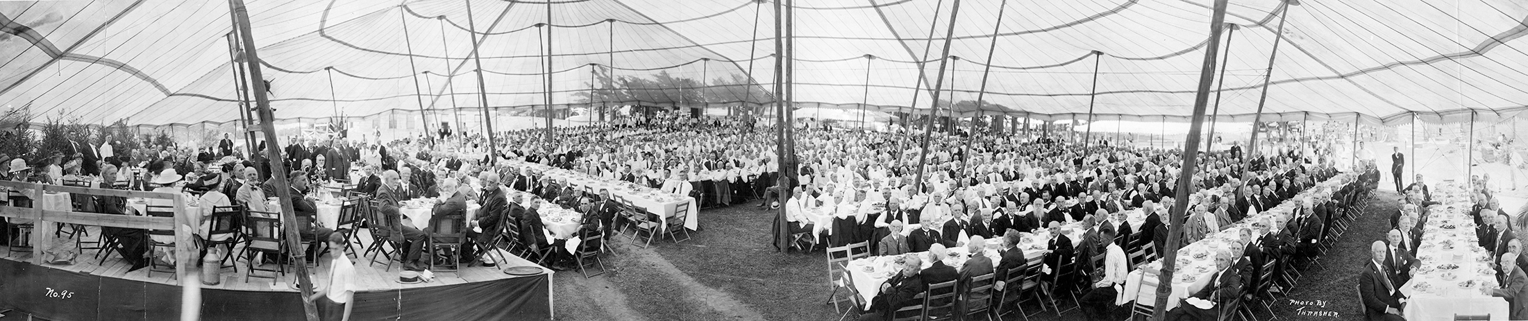 A wide black and white photo of hundreds of people eating under a large pent
