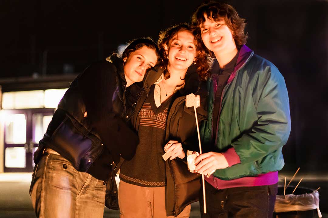 Three student, leaning on one another, hold up a roasted marshmallow.