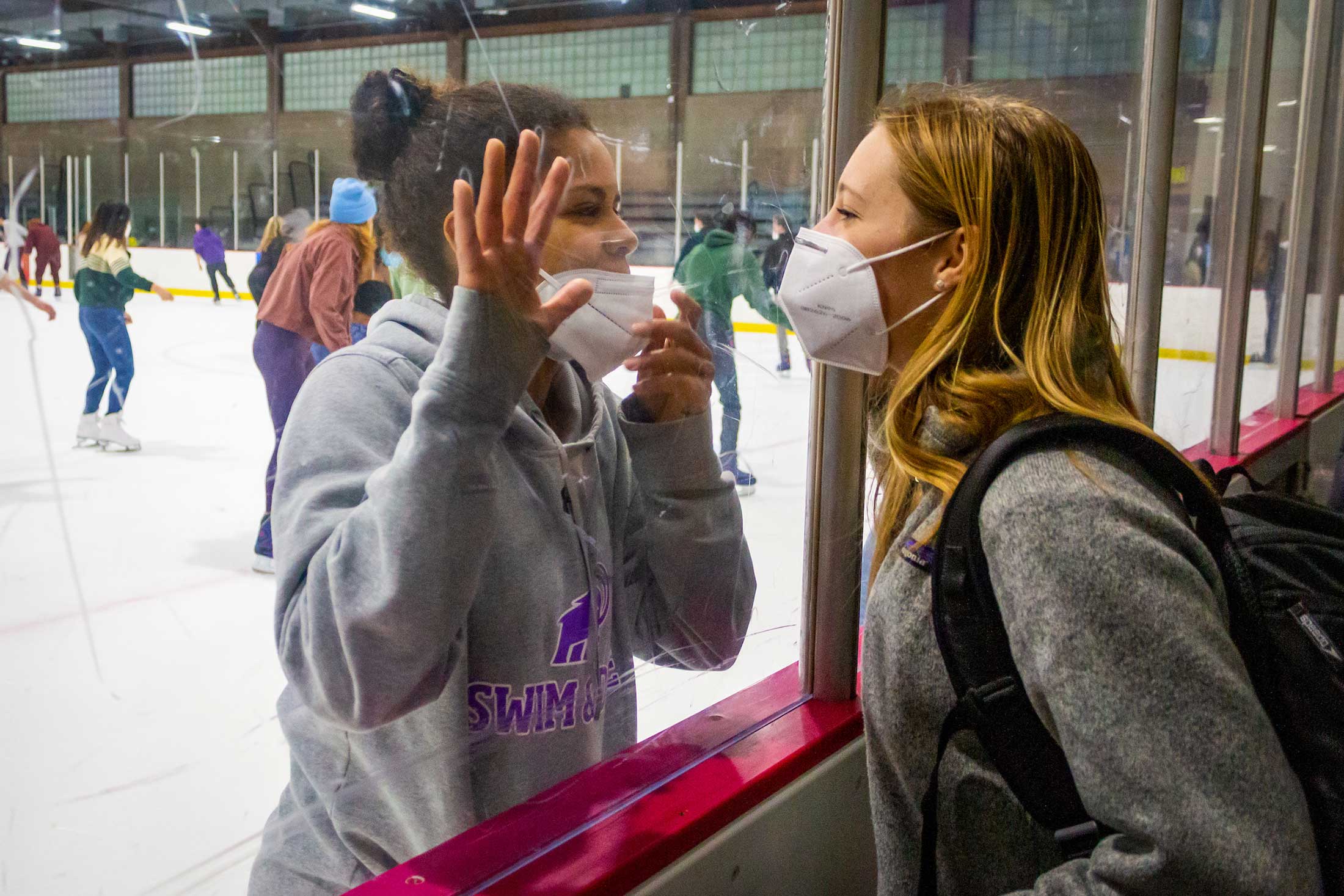 Two masked students converse from opposite side of a plexiglass divider at the Orr Rink.