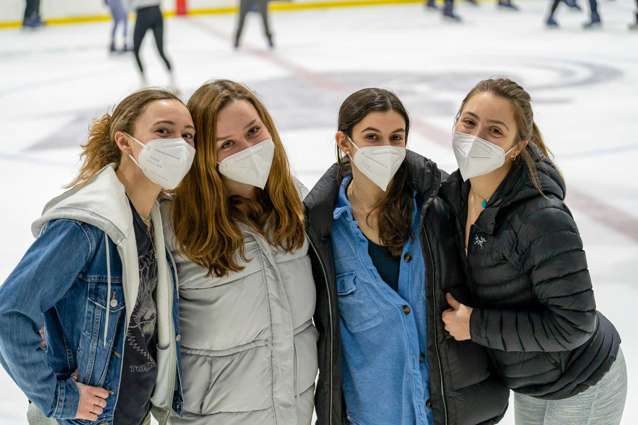 Four female students, all wearing face masks, stop to pose for a photo while ice skating.
