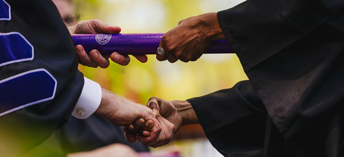 A close of two people shaking hands while exchanging a diplioma in a purple tube. 