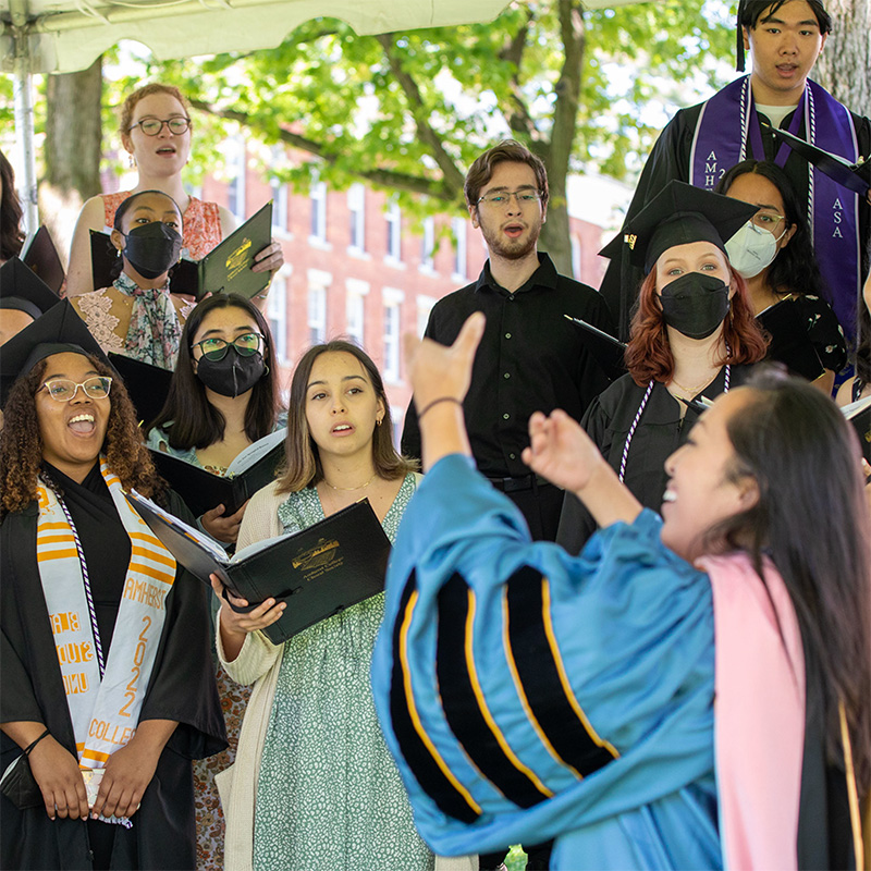 A choir singing outside led by a woman in commencement regalia