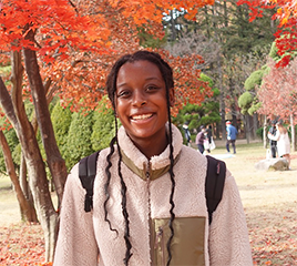 A young woman, smiling, in a beige jacket, poses in front of trees full of red and orange leaves.