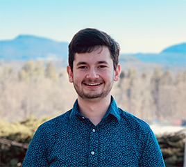 A young man in a blue shirt, smiling, on a sunny day, with the Holyoke Range visible behind him.