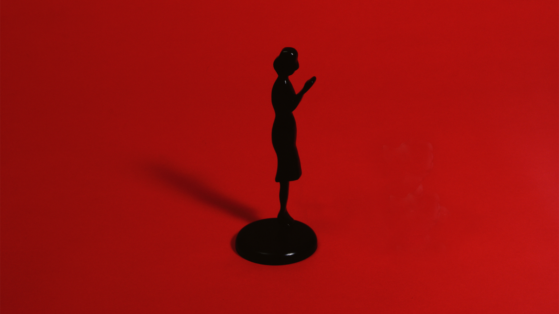  A silhouette figurine in a dress is set on a red background. It casts a shadow, and its arms are bent upward at the elbow.