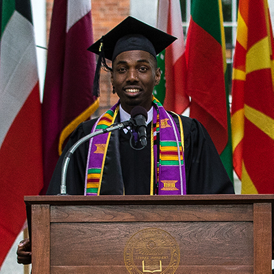 A young man wearing a cap and gown speaking at commencement