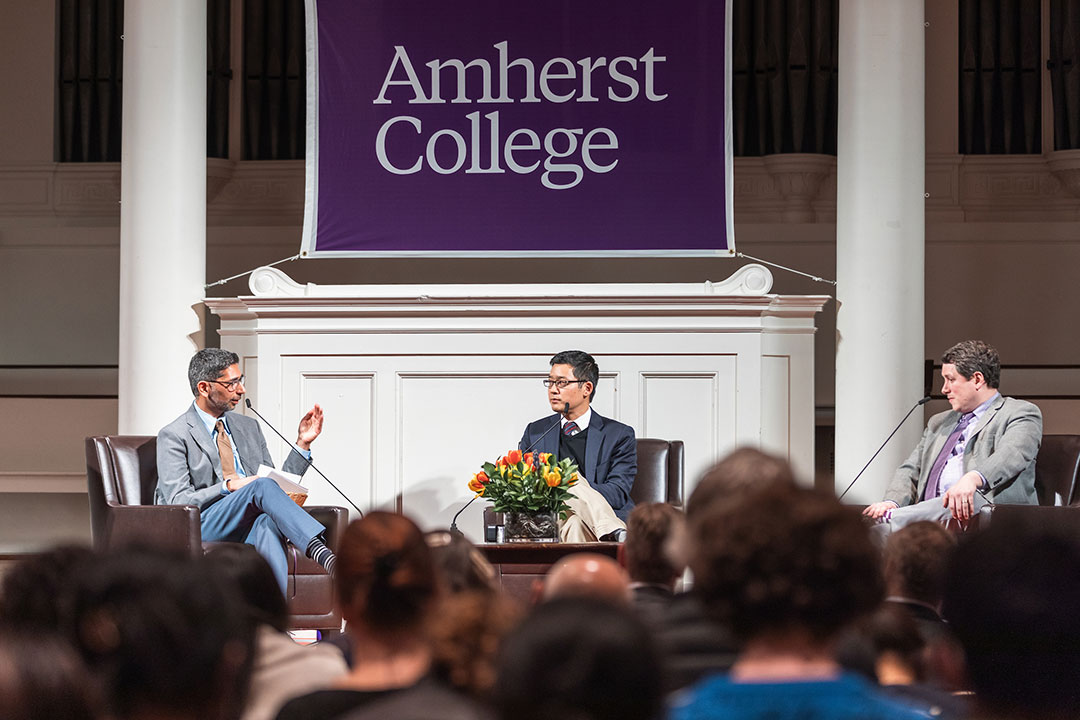 Three men in suits sitting on a stage with an Amherst College banner hanging behind them.