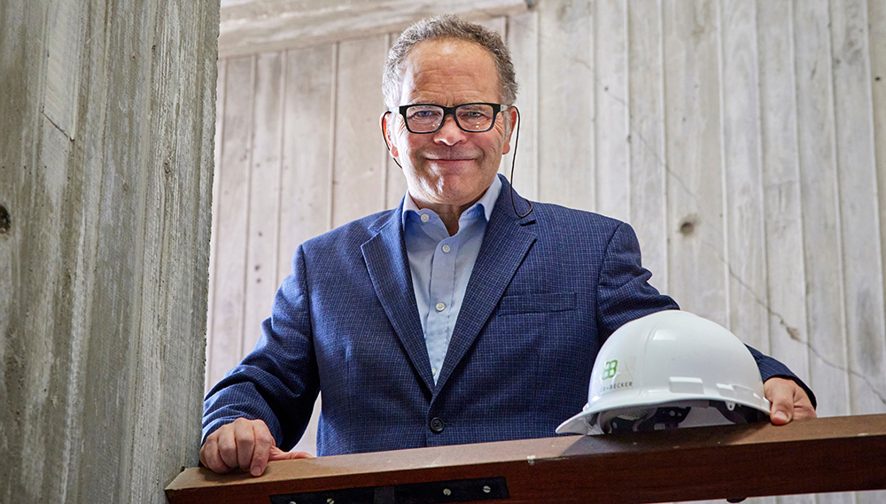A man with short gray hair and glasses holding a hard hat