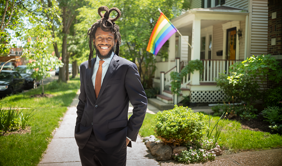 A Black man in a suit standing in a suburban neighborhood with a Pride flag in the background