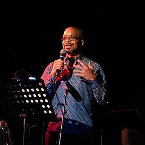 A Black man staging on  dark stage speaking into a microphone