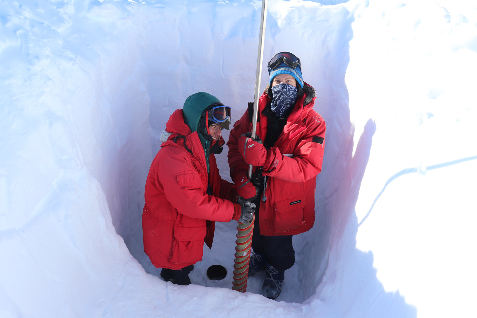 two people in heavy parkas stand in a hole in the snow holding a pole with a drill to extract ice