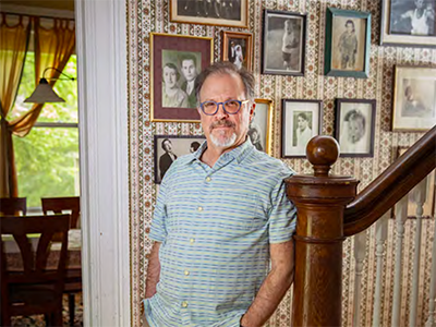 A man standing in a nice home leaning against a stairway railing with family photos in the background
