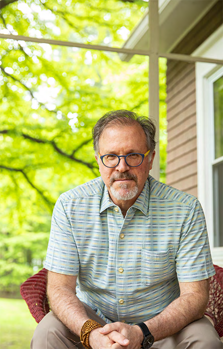 A man in glasses sitting on a porch surrounded by green trees