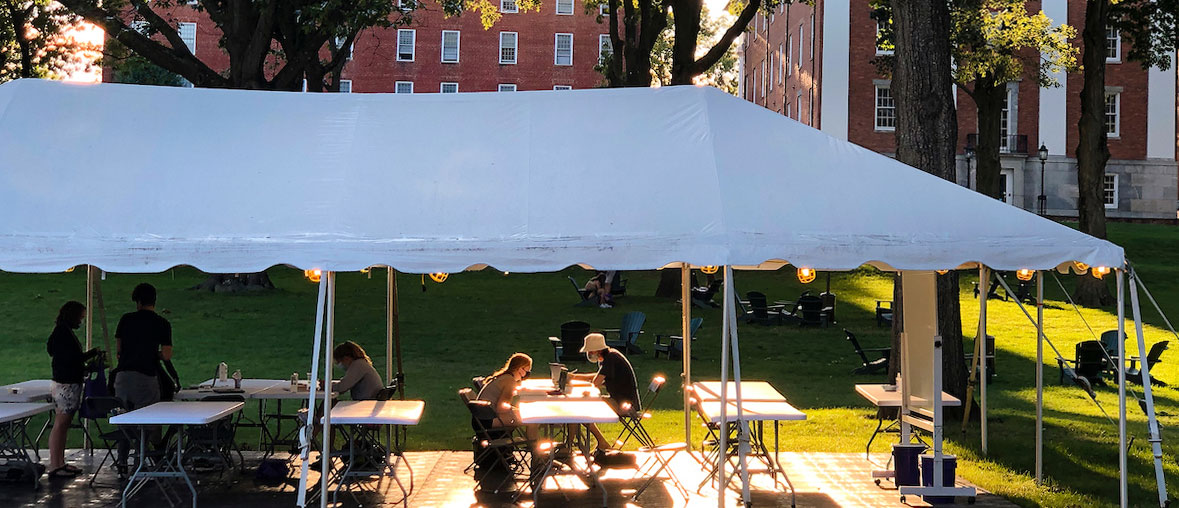 students wearing masks study outside under a tent together with sunlight streaming onto the quad
