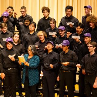 Choral Society singers standing on risers on the Buckley stage; most are dressed in black, and some are wearing purple Amherst College caps