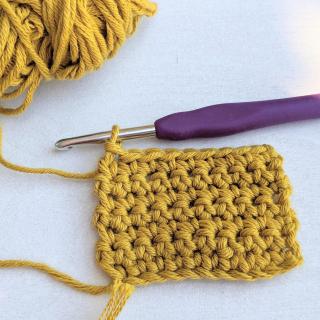 A square made of dark yellow crochet yarn, still looped on a crochet hook with a purple handle