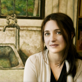 Simone Dinnerstein looks into the camera, wearing a white sweater and black V-neck. Behind her are a painting of a sink and window with a green world outside.