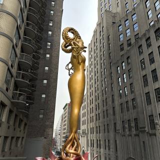 Large gold statue standing on a red flower, between tall buildings