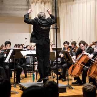 Mark Swanson is shown hopping an inch or two into the air above his podium as he conducts the orchestra, arms raised, back to audience. The strings section around him is wearing concert black, holding bows to instruments.