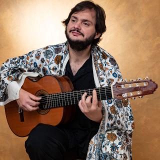 Man with beard holding guitar, wearing black with colorful shawl 
