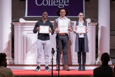 Three students standing on a stage in front of an Amherst College banner, holding certificates.