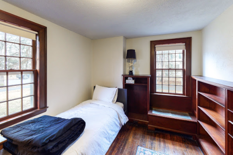 Image of a white twin-sized bed with a black blanket at the foot. On two walls are brown-framed windows, and on the third visible wall is a wooden bookshelf.