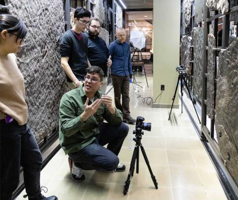 Tim Pinault kneels in front of a camera and explains to nearby students how to take photographs of adjacent fossil-laden stone slabs from different angles.