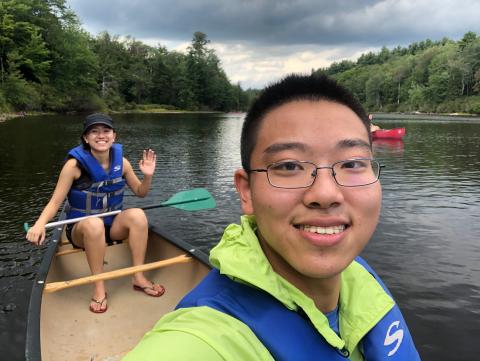 My friend and I are sitting on a canoe on a lake. My friend is taking the selfie and I am waving.