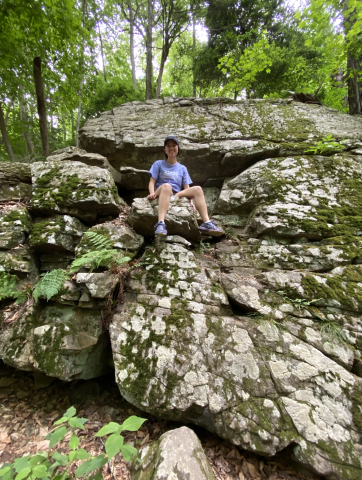 Sitting on a large rock at the park in new jersey, in t-shirt, smiling down at camera